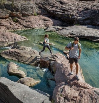 Two people hike across hill country rock to cross a clear flowing stream.